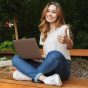 Smiling young girl using laptop computer while sitting on a bench outdoors and showing thumbs up